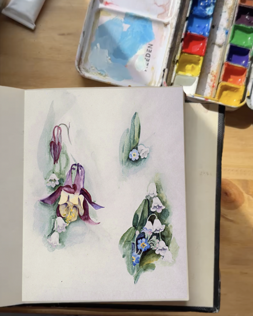 Photograph of a books open to a page with small paintings of flowers in watercolor. In the top-right corner is a small watercolor palette.